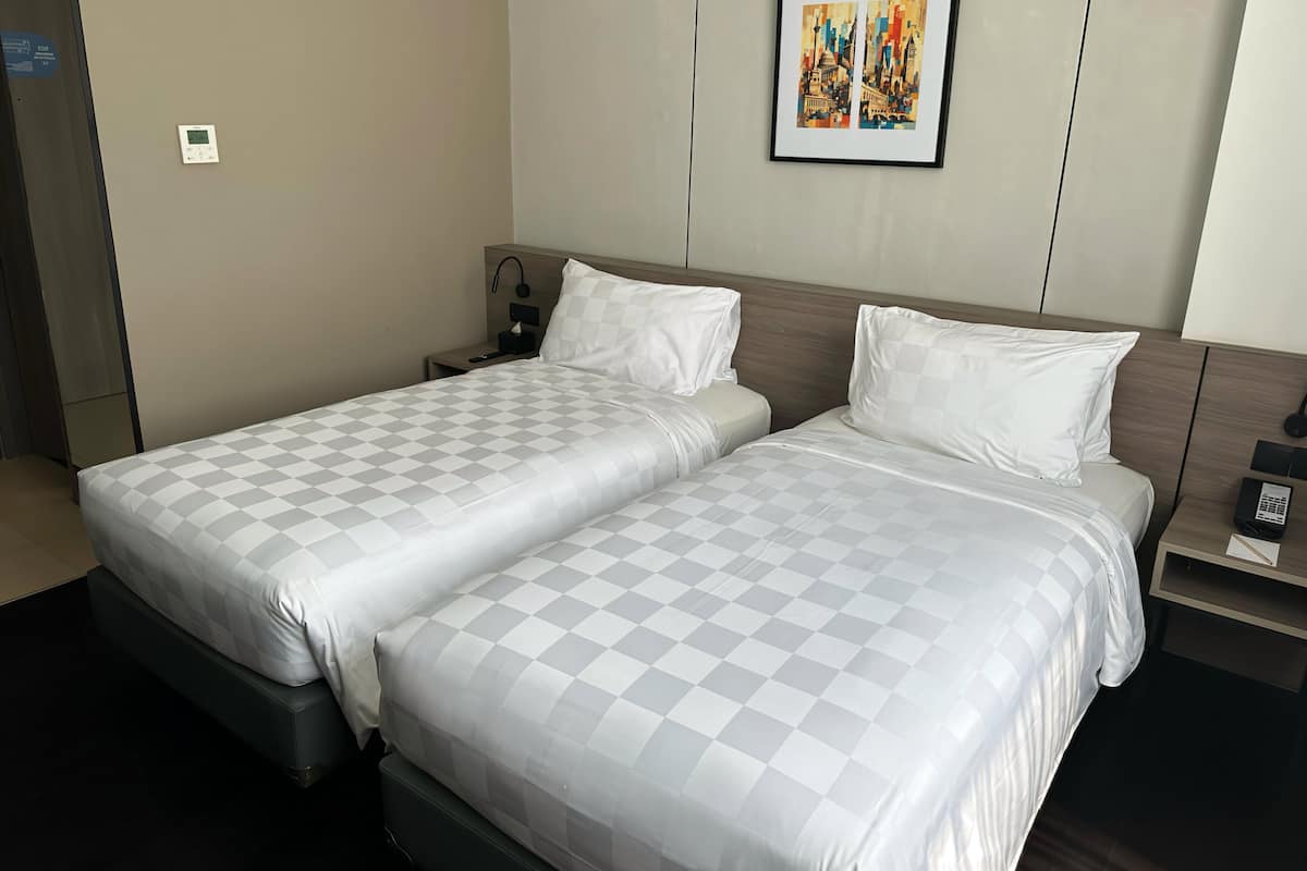 Une chambre d’hôtel avec deux lits, une table de chevet, une commode, un thermostat fixé au mur et une œuvre d’art encadrée au-dessus des lits.