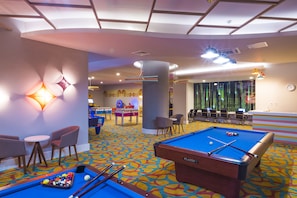 Une table de billard recouverte d’un drap bleu, un queue de billard et plusieurs billes. Il y a deux chaises et une petite table avec un néon fixé au mur.
