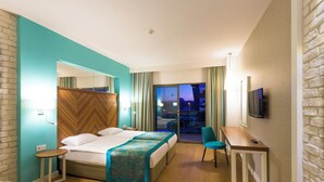 A hotel room with a bed, a desk, a chair, a TV, and a view of the city at night. - Terrace Elite Resort - All Inclusive (Manavgat)