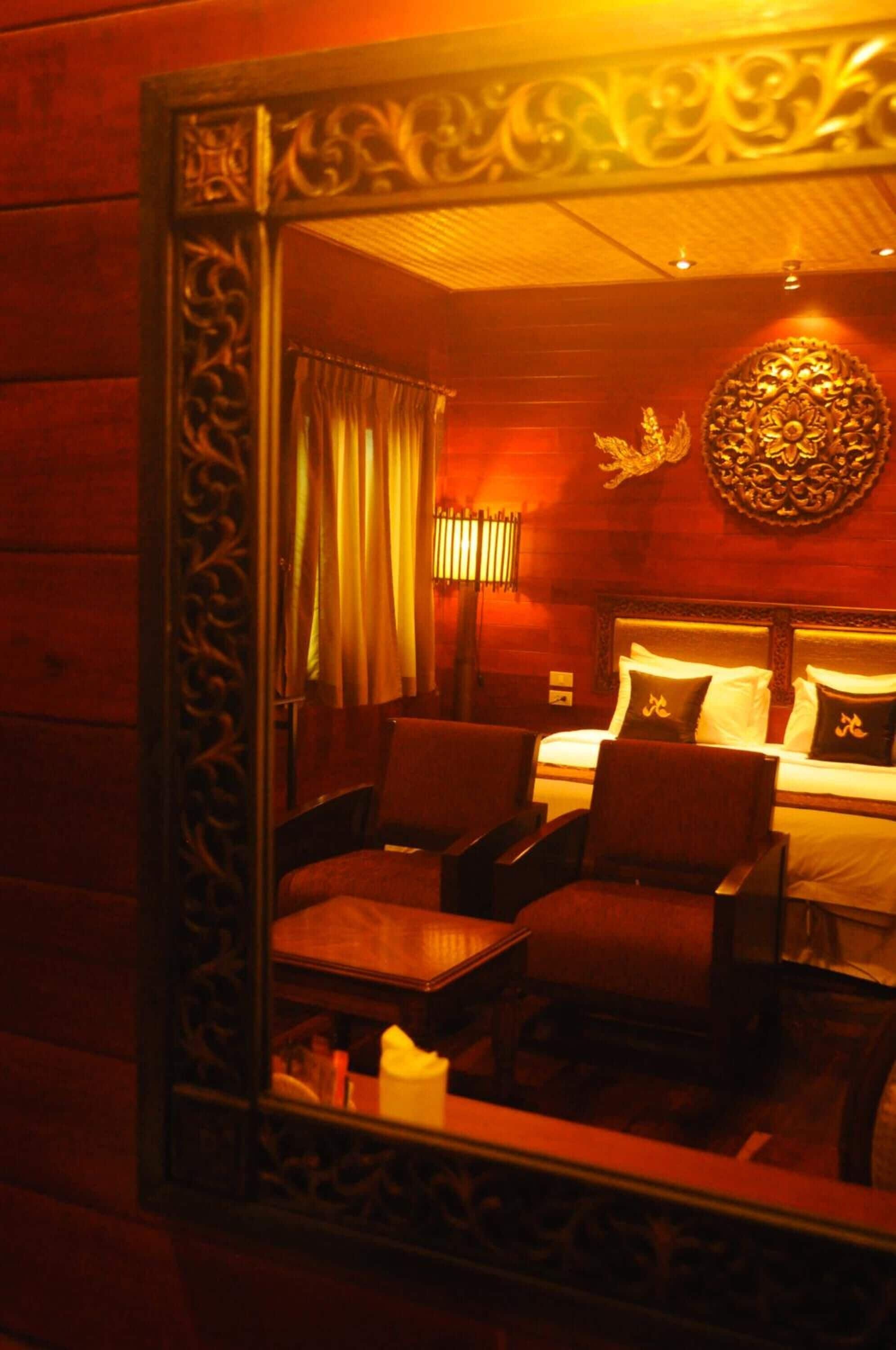A room with a bed, two chairs, a table, and a mirror reflecting the room.