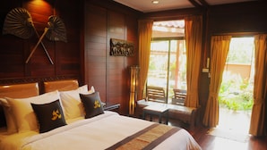 A hotel room with a large bed, a wooden headboard, a small table, and a view of the outdoors through a sliding door. - Ruen Kanok Thai House (Hua Hin)