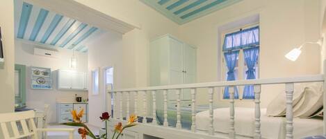 Una habitación con una cama blanca, una silla blanca, una mesita con un jarrón de flores y una ventana con cortinas azules.