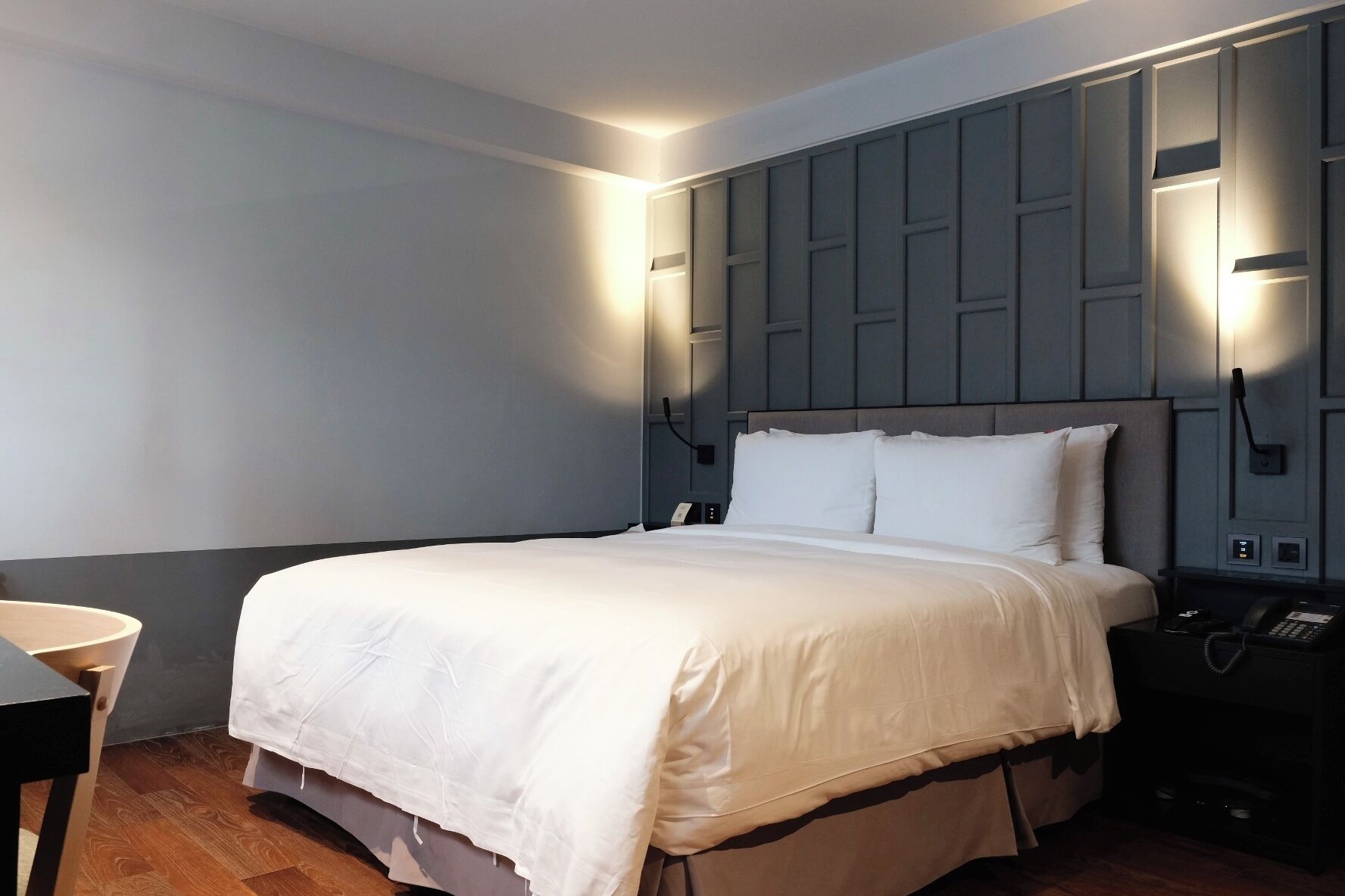 A neatly made bed with white linens, a bedside table with a phone, and a wall-mounted light fixture.