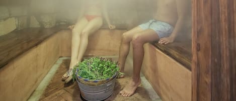 Una sauna con dos personas sentadas en bancos, un cubo de plantas y paredes de madera.