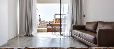 Ein Wohnzimmer mit braunem Ledersofa, einer farbenfrohen Stickdecke und Blick auf eine Stadtlandschaft durch eine Glastür.