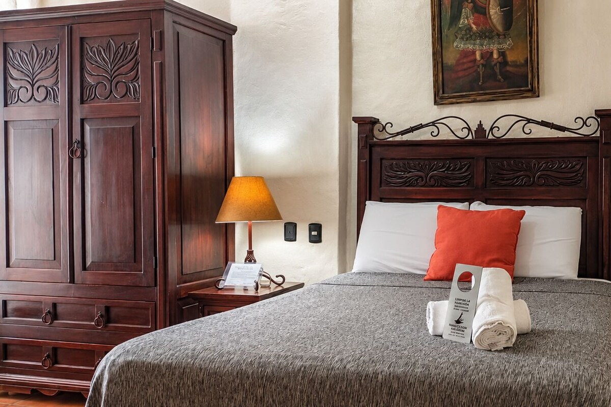Habitación de hotel con un armario de madera, una cama con cubrecama gris, almohada roja y toalla blanca sobre la cama.