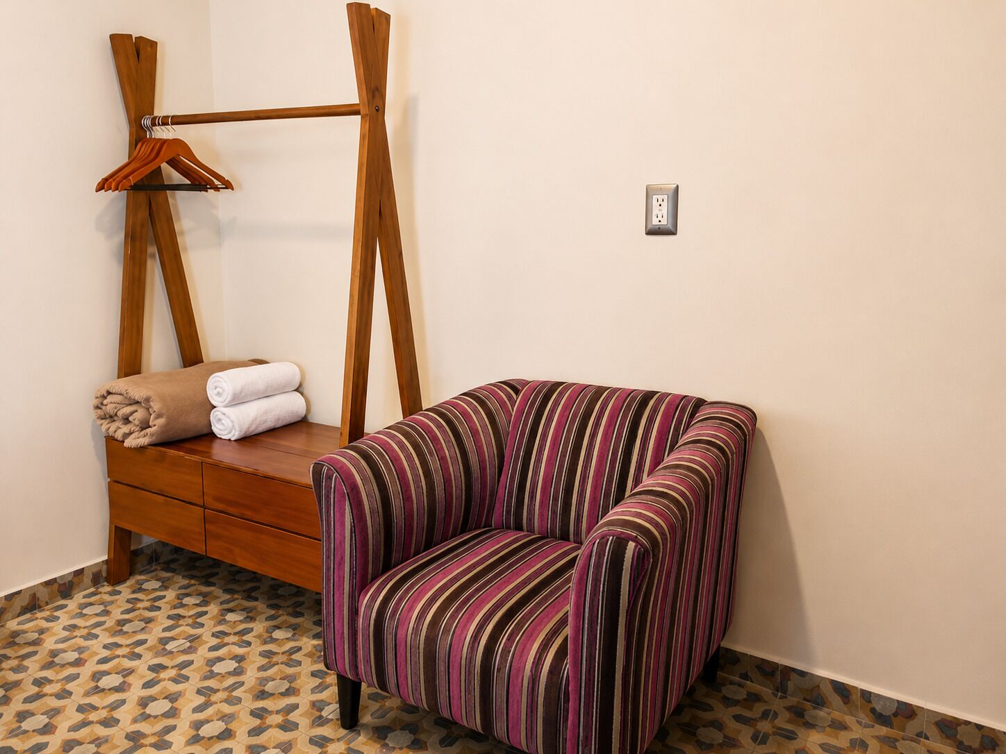 Una habitación con un sillón a rayas, un banco de madera con toallas y un perchero de madera en la pared.