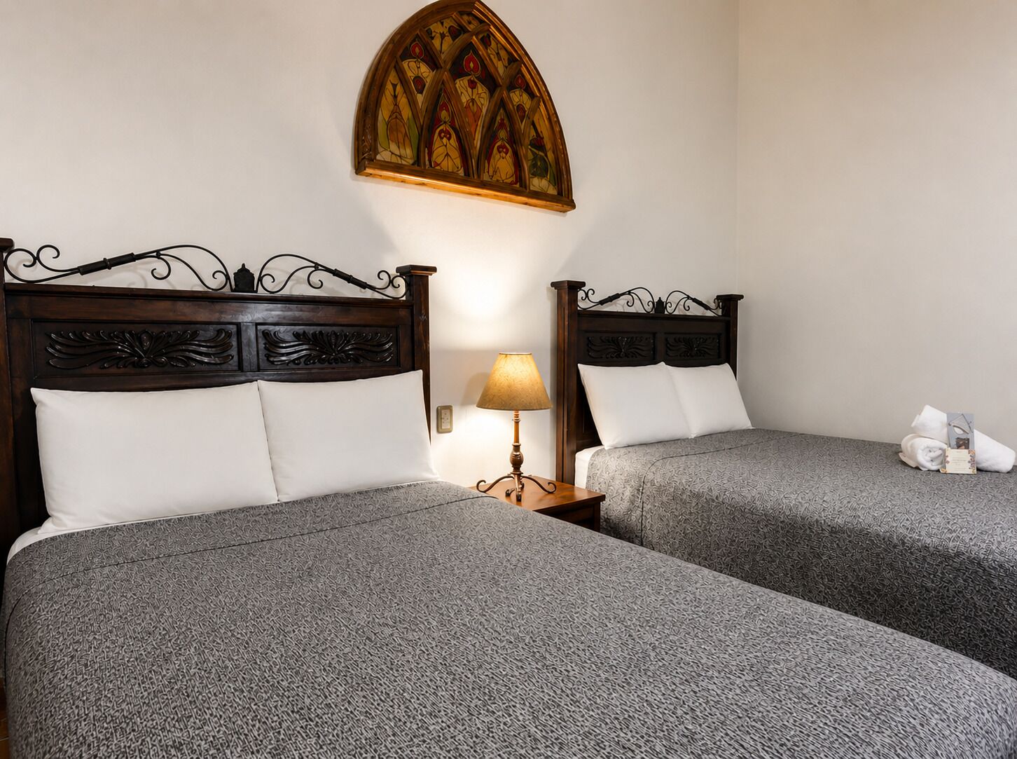 Quarto de hotel com duas camas, uma mesa de cabeceira com abajur e um objeto decorativo na parede acima das camas.