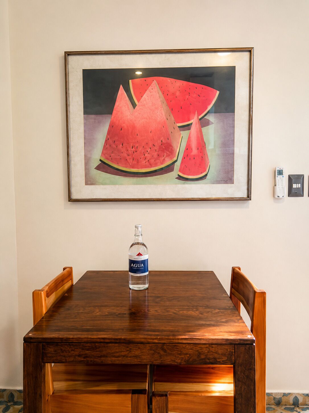 A dining table with a bottle of Agua on it, framed artwork above, and a wall socket to the right.