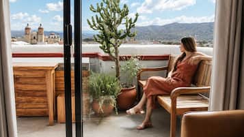 Una persona sentada en un banco de madera en un balcón con plantas en macetas y vista a un edificio con cúpulas.