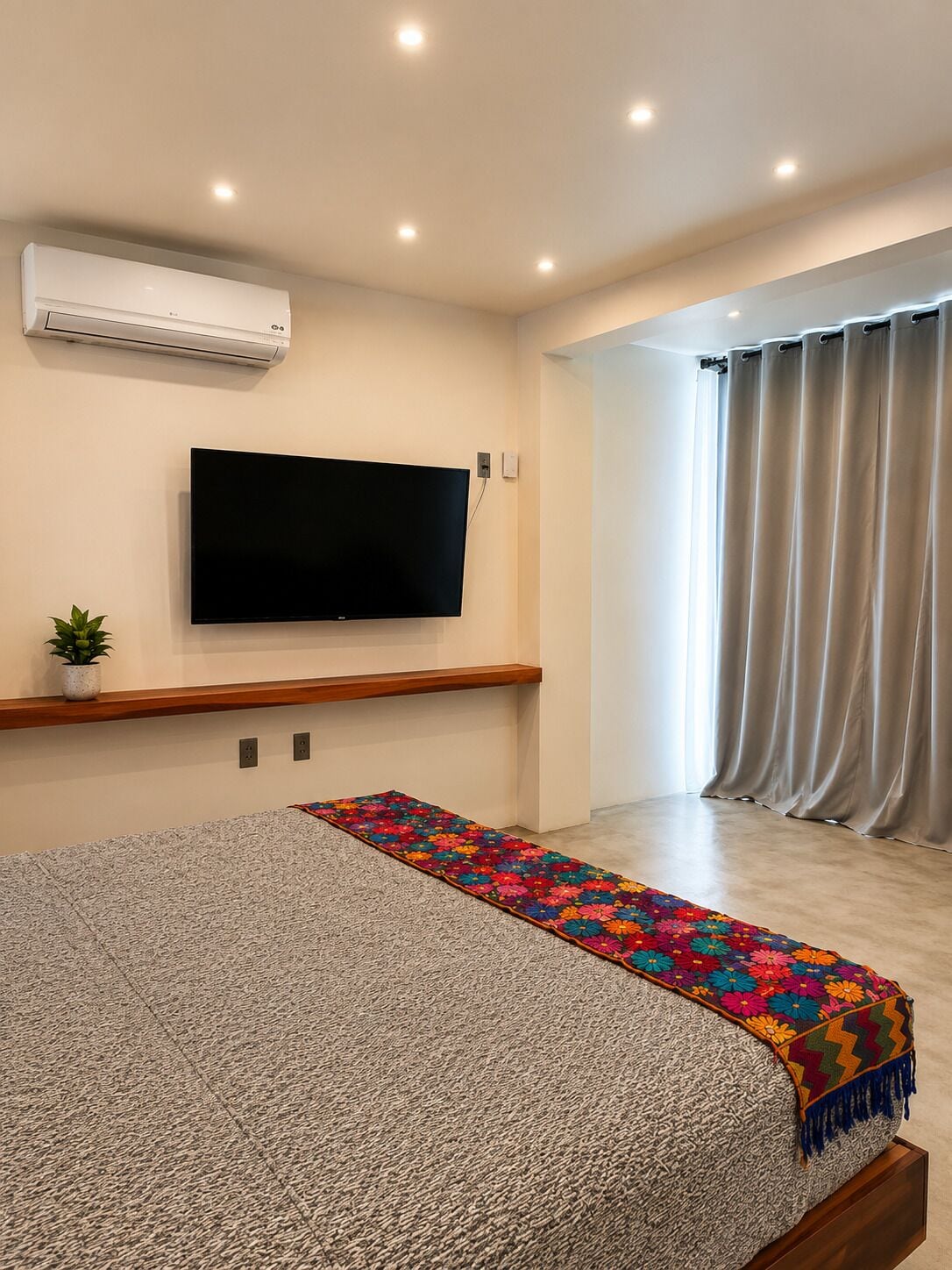 A bedroom with a flat-screen TV mounted on the wall, a wooden shelf below, a patterned bedspread, a small potted plant, and a large window with curtains.