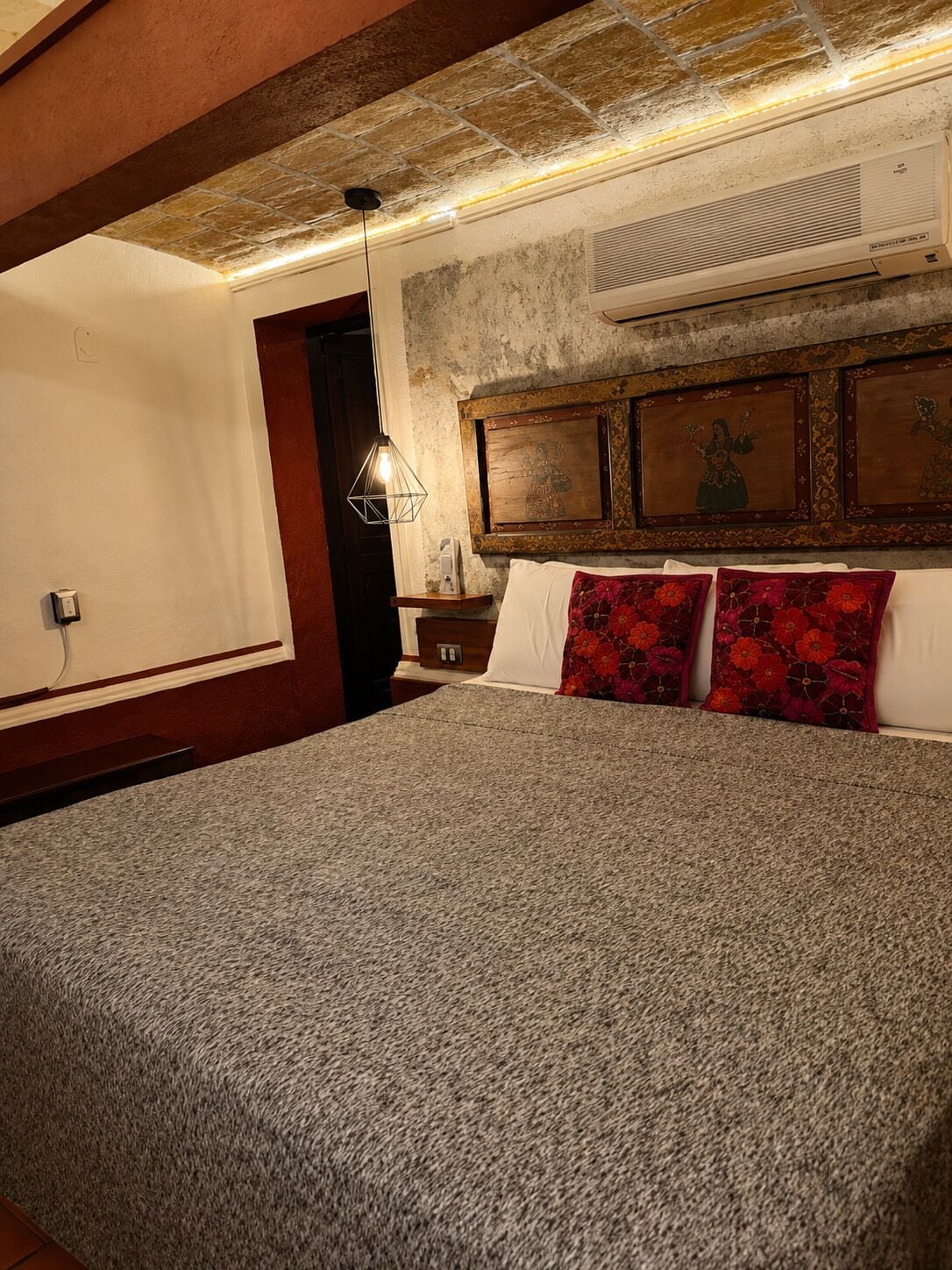 A bedroom with a large bed, a nightstand, a wall-mounted heater, and a wooden headboard with artwork.
