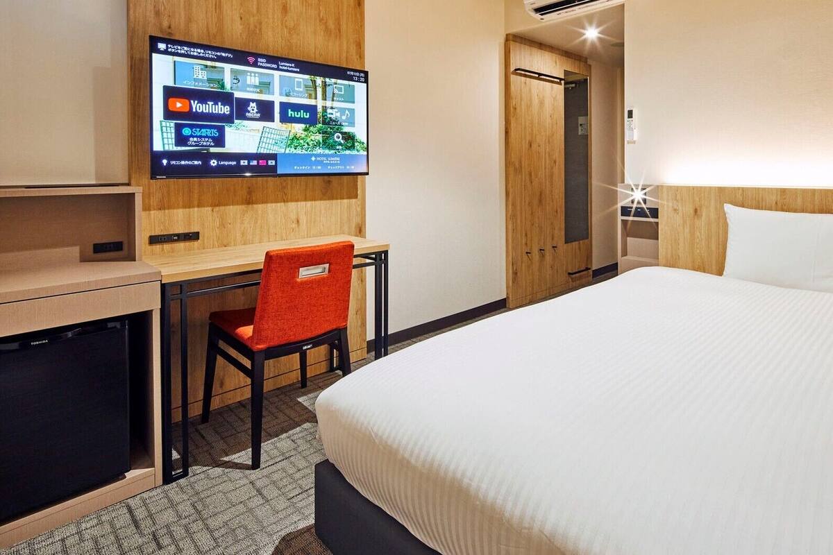 A hotel room with a bed, a desk with a chair, a TV displaying streaming services, and a wooden wardrobe.