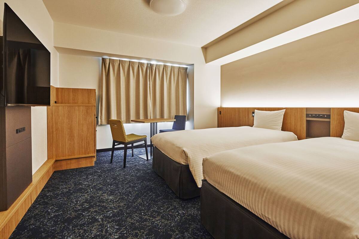 A hotel room with two beds, a desk, a chair, a television, and a window with curtains.