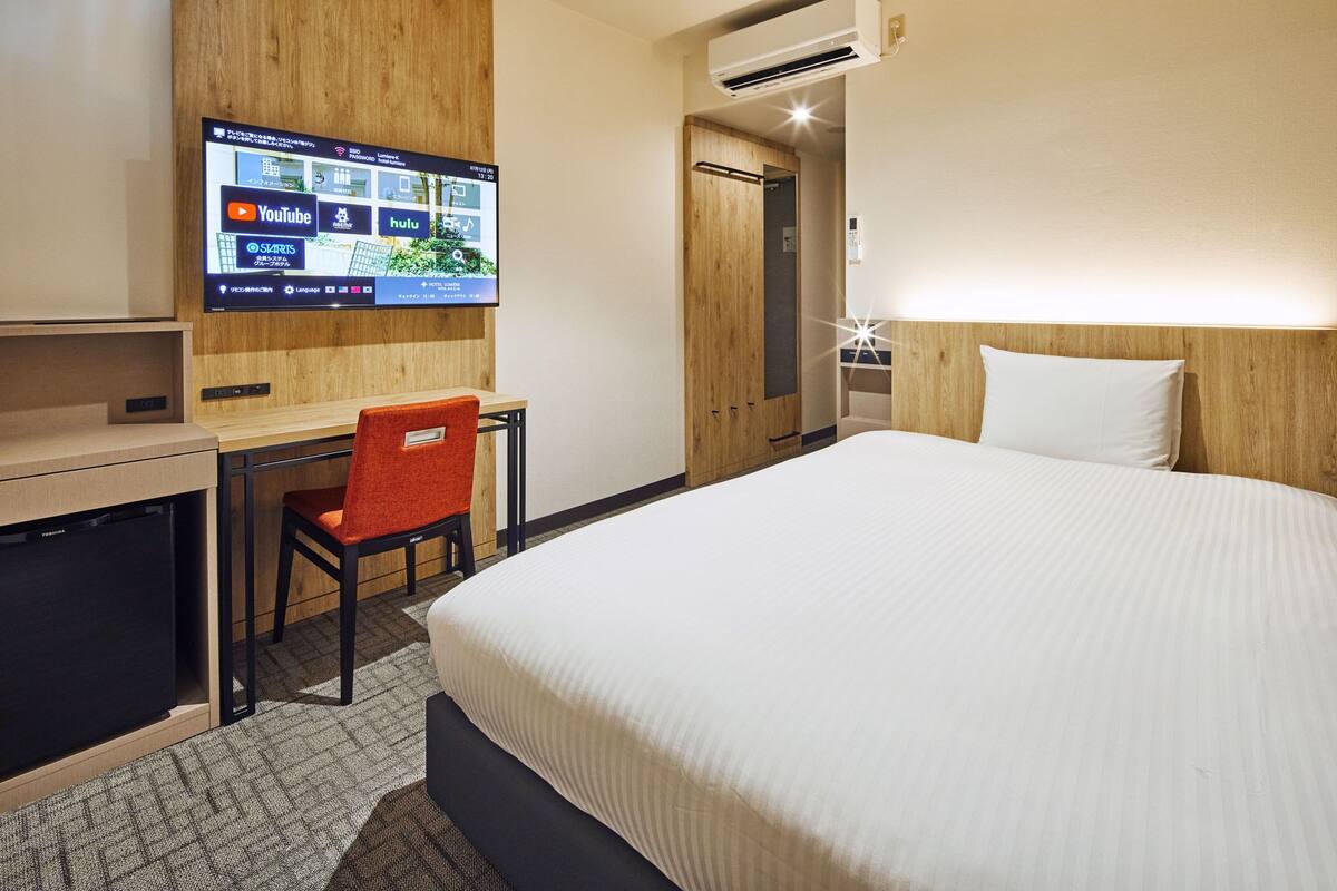 A hotel room with a bed, a desk with a chair, a television mounted on the wall, and an air conditioning unit.