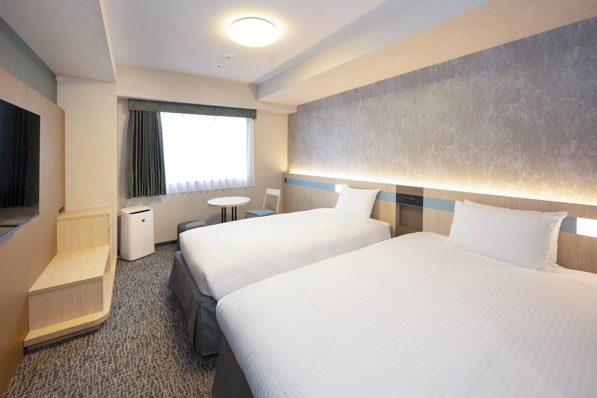 Une chambre d’hôtel avec deux lits, un bureau, une chaise, une petite table et une fenêtre avec des rideaux.