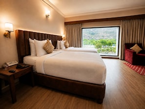 Habitación de hotel con dos camas, un cabecero de madera, una mesita de noche con teléfono y control remoto, y vista a zonas verdes por la ventana.