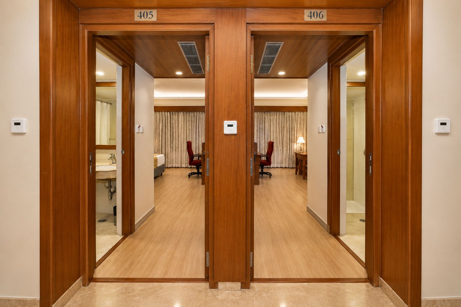 A hotel room with a wooden door, a bed, a desk, a chair, a lamp, and a bathroom with a sink and mirror.