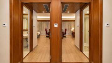 A hotel room with a wooden door, a bed, a desk, a chair, a lamp, and a bathroom with a sink and mirror.