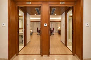 Habitación de hotel con puerta de madera, cama, escritorio, silla, lámpara y baño con lavamanos y espejo.