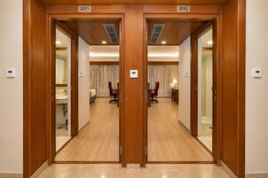Quarto de hotel com porta de madeira, cama, escrivaninha, cadeira, luminária e banheiro com pia e espelho.