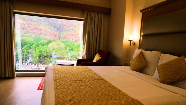 A hotel room with a large bed, a view of a mountainous landscape, a small table, and a chair.