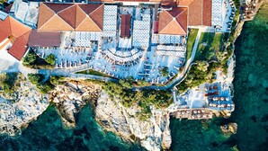 An aerial view of a coastal hotel with multiple dining areas, outdoor seating, and a rocky shoreline.