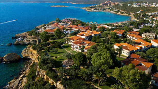 An aerial view of a coastal town with red-roofed buildings, a marina, and wind turbines in the distance.