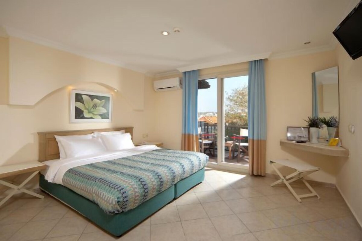 A hotel room with a bed, a desk, a chair, a window with curtains, and a plant on a shelf.