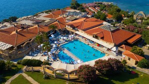 An aerial view of a coastal hotel with a large swimming pool, multiple sun loungers, and a view of the sea and wind turbines in the background.