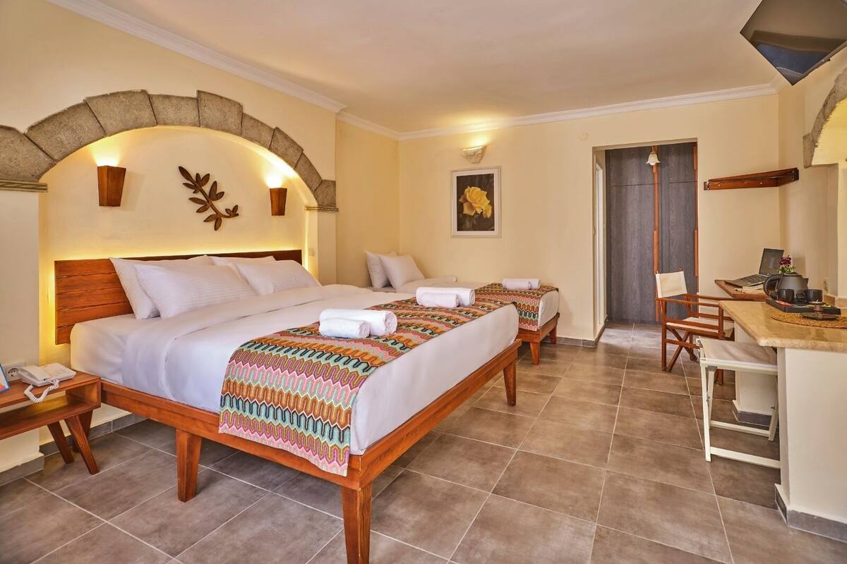A hotel room with a large bed, two smaller beds, a desk with a chair, and a bathroom visible through an archway.