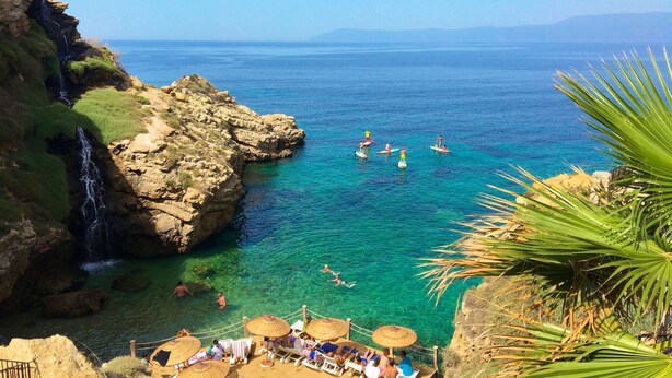 A coastal area with clear waters, a rocky cliff, and a beach with umbrellas and lounge chairs.
