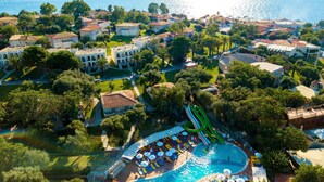 An aerial view of a resort with a central swimming pool, multiple buildings, and a view of the sea.