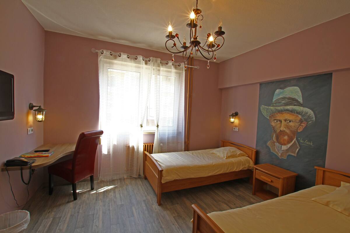 A bedroom with a wooden bed, a desk with a chair, a chandelier, and a painting on the wall.