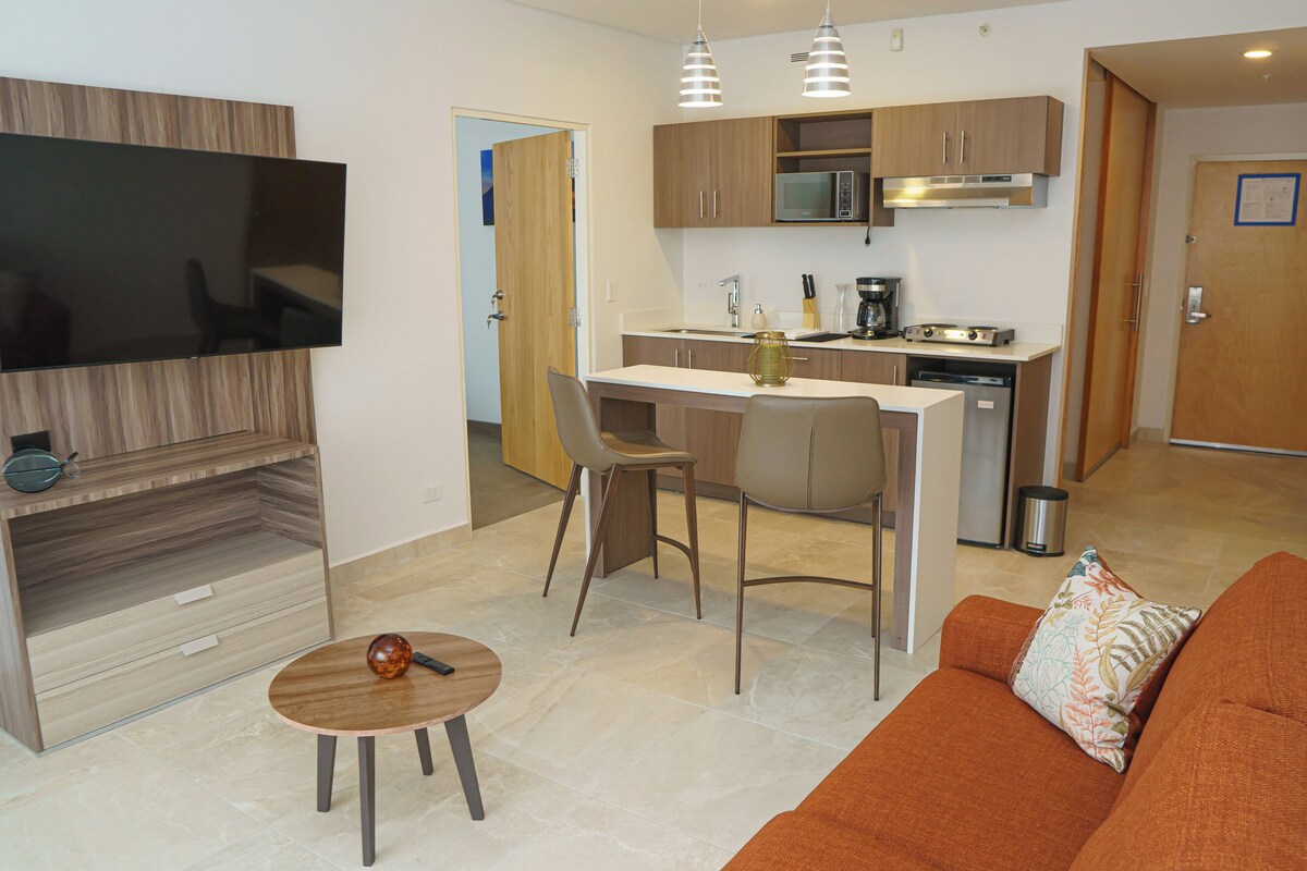 A modern kitchen with a microwave, stove, and sink. There is a dining area with a table and chairs. A flat-screen TV is mounted on the wall.