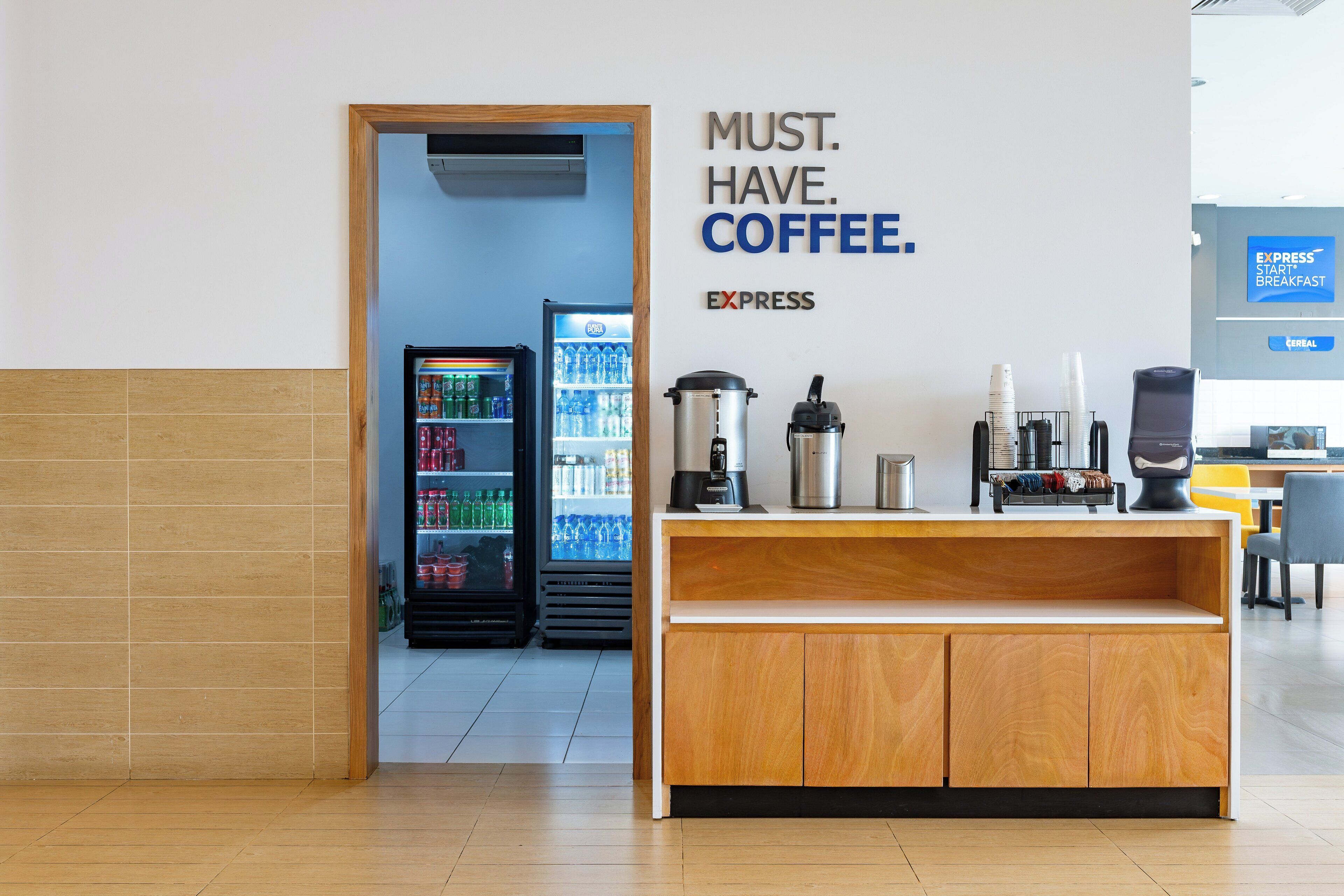 Una estación de café con un cartel que dice "MUST. HAVE. COFFEE." y un refrigerador lleno de bebidas.