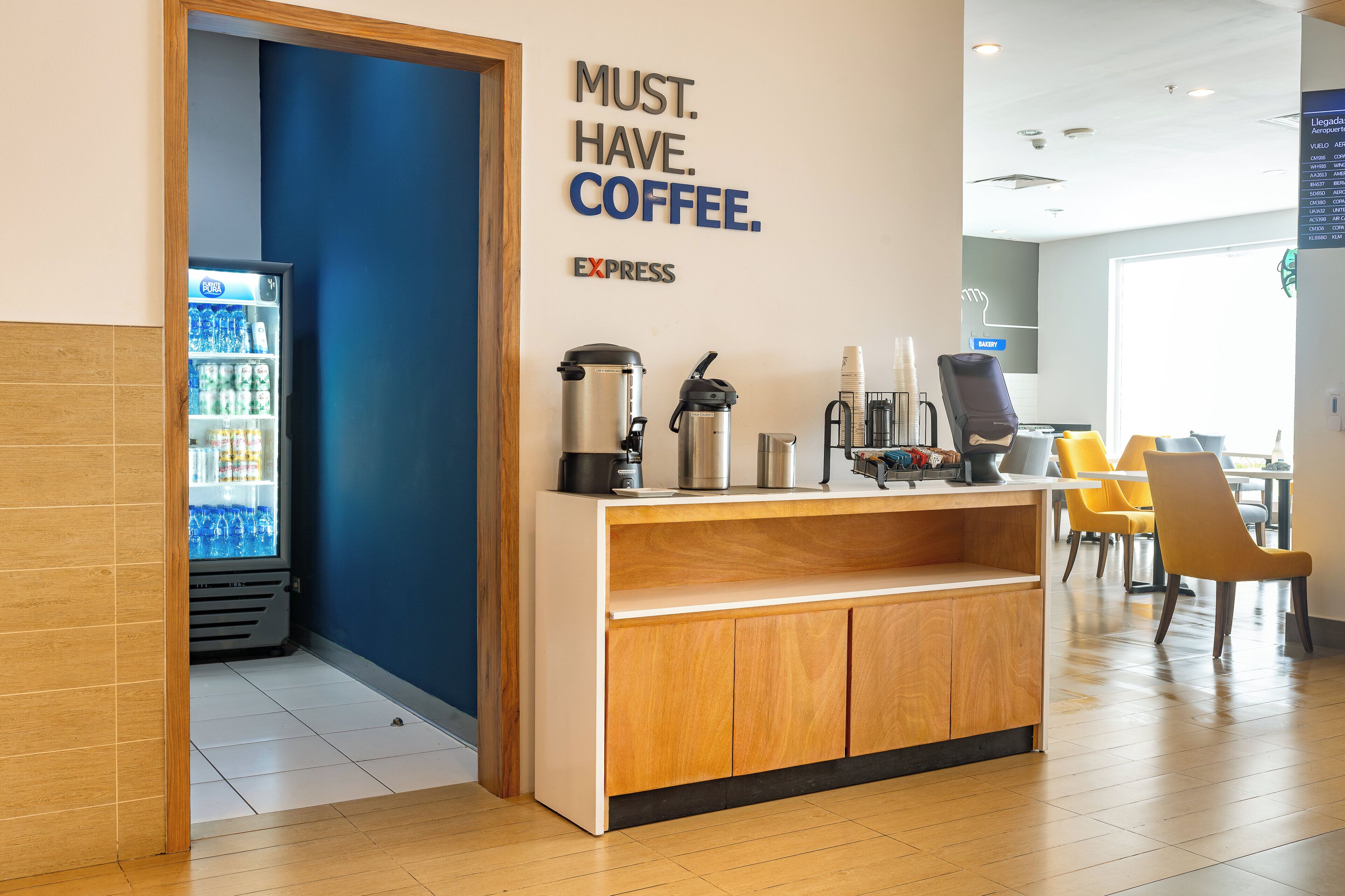 A coffee station with a sign that reads "MUST. HAVE. COFFEE." and a refrigerator stocked with beverages.