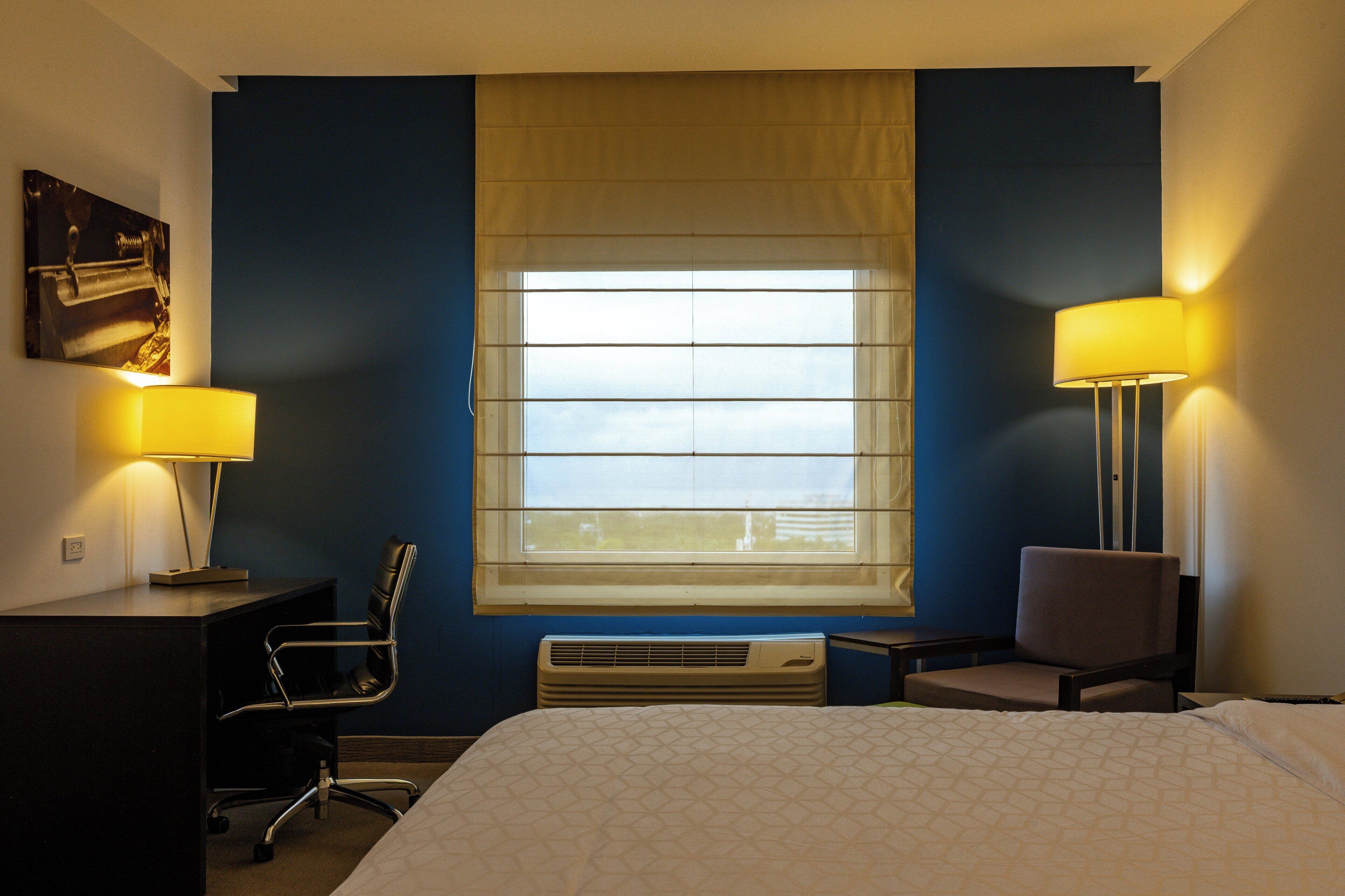 Une chambre d’hôtel avec un lit, deux chaises, un bureau, une lampe et une fenêtre avec des stores.