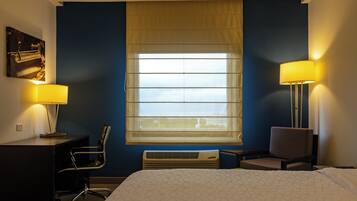 A hotel room with a bed, two chairs, a desk, a lamp, and a window with blinds.