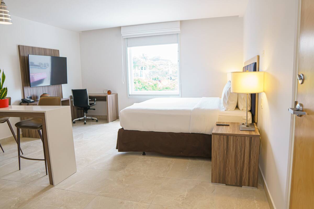 A hotel room with a bed, desk, chair, TV, lamp, and a window with a view.