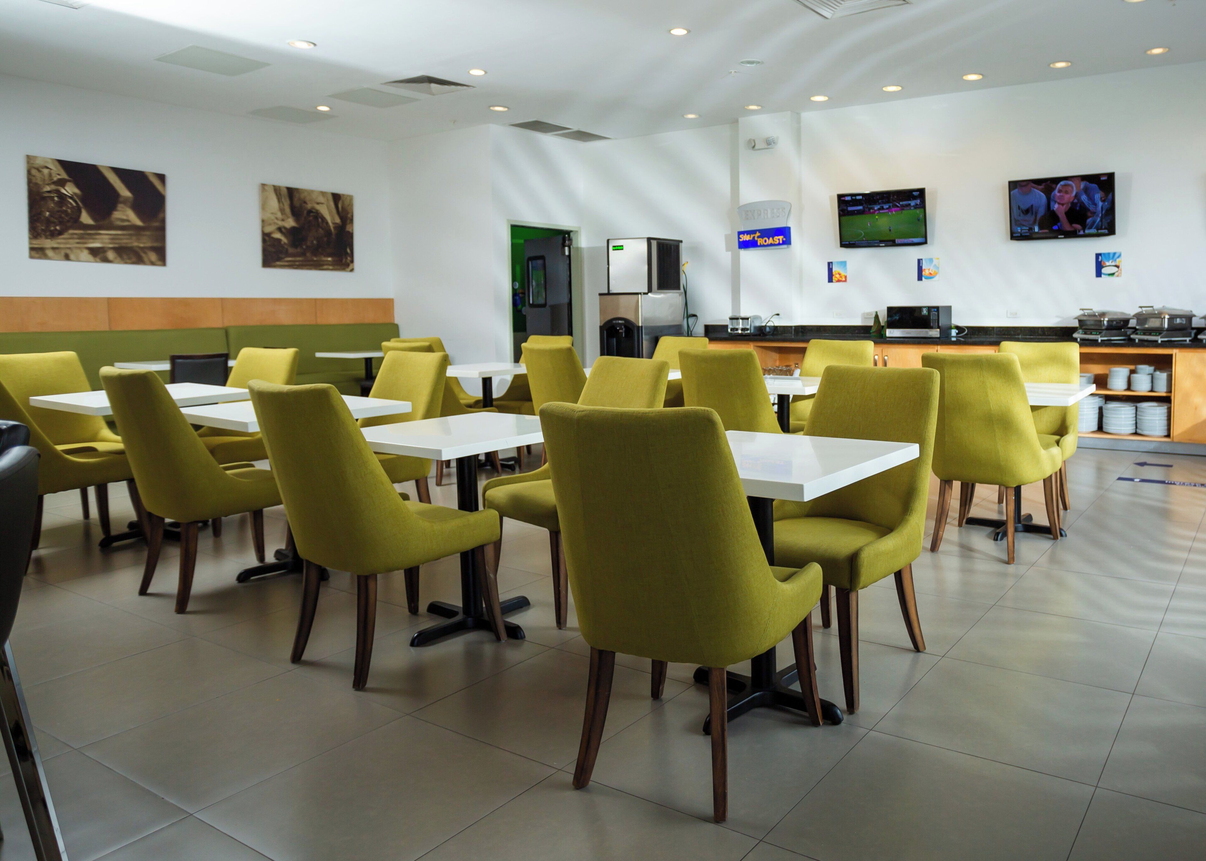 A restaurant with yellow chairs and white tables, a kitchen area, and a television screen.