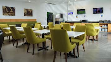 A restaurant with yellow chairs and white tables, a kitchen area, and a television screen.