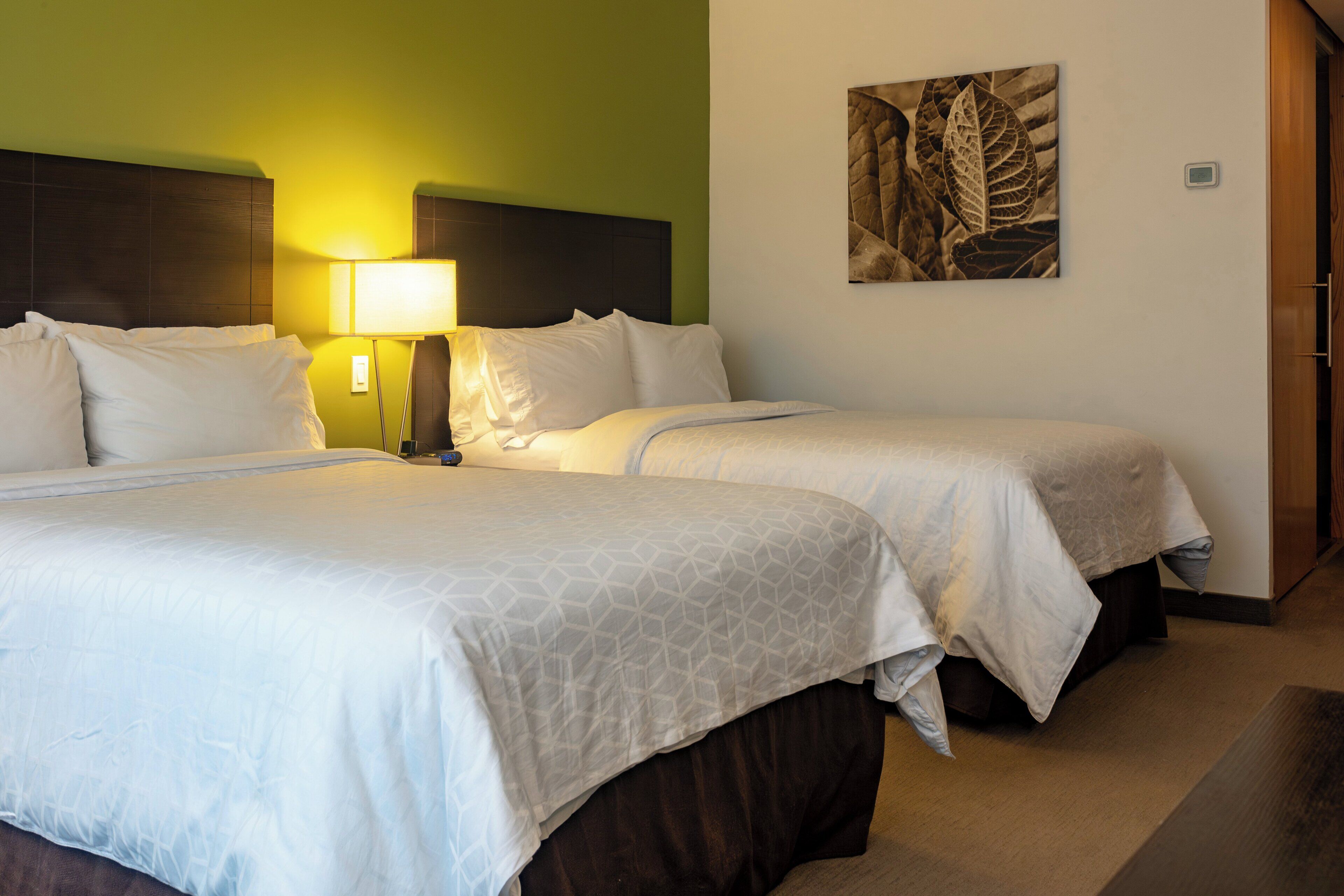 Habitación de hotel con dos camas individuales, una mesita de noche con lámpara y una obra de arte colgada en la pared.