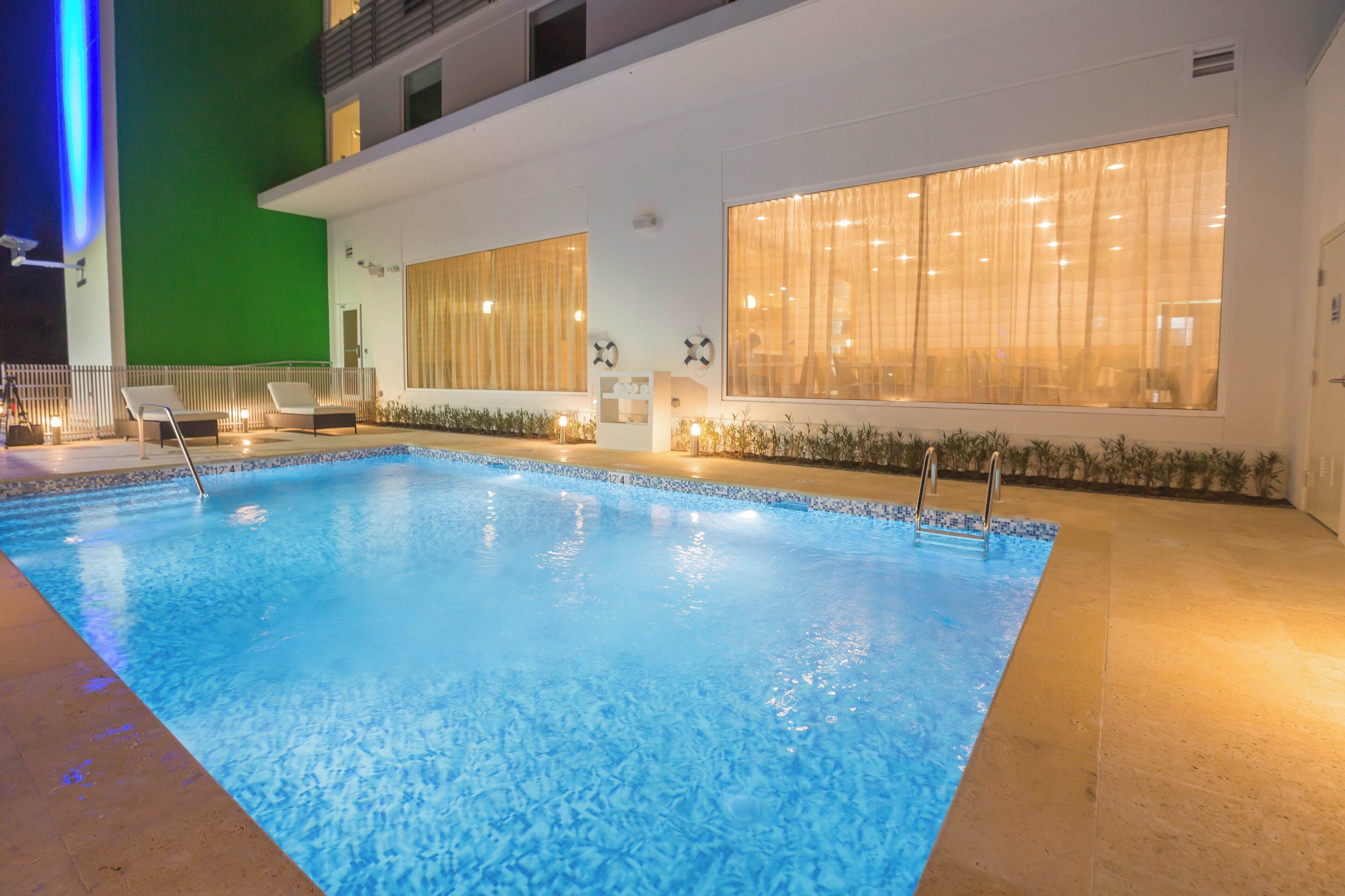 A swimming pool with a tiled edge, a lounge chair, and a window with curtains.