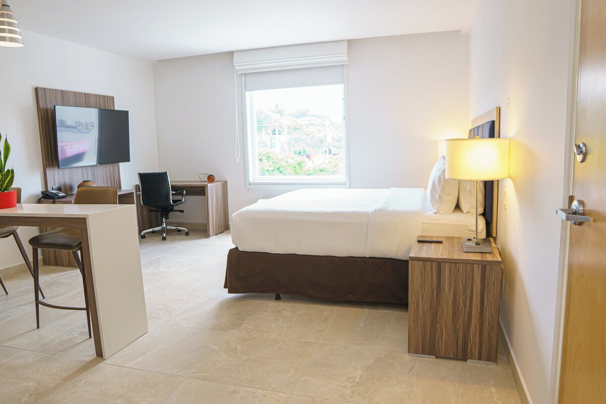 A hotel room with a bed, desk, chair, TV, lamp, and a window with a view.