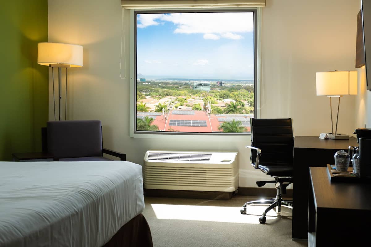 A hotel room with a bed, desk, chair, and a large window offering a city view.