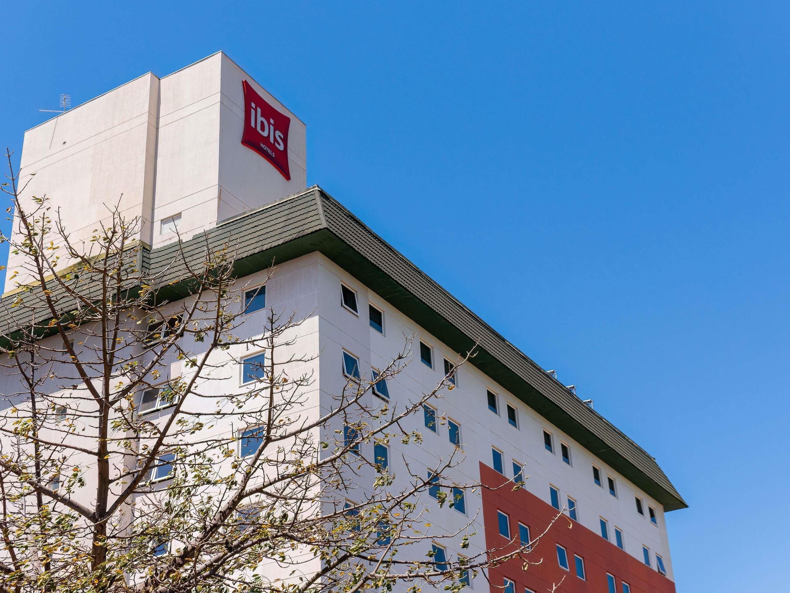 Edifício de hotel com logotipo vermelho e branco bem visível, várias janelas e céu azul.