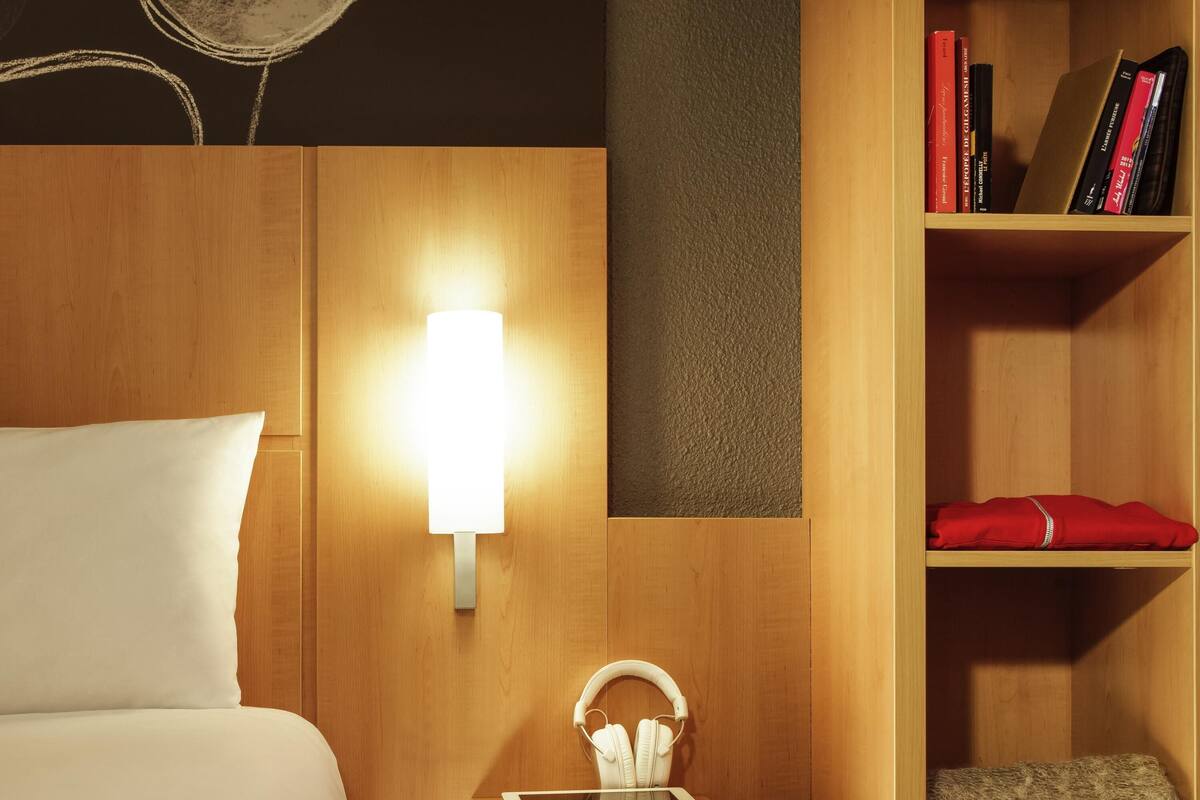 Une chambre d’hôtel comprenant un lit, une table de chevet avec une tablette et des écouteurs, une lampe fixée au mur et une bibliothèque remplie de livres.