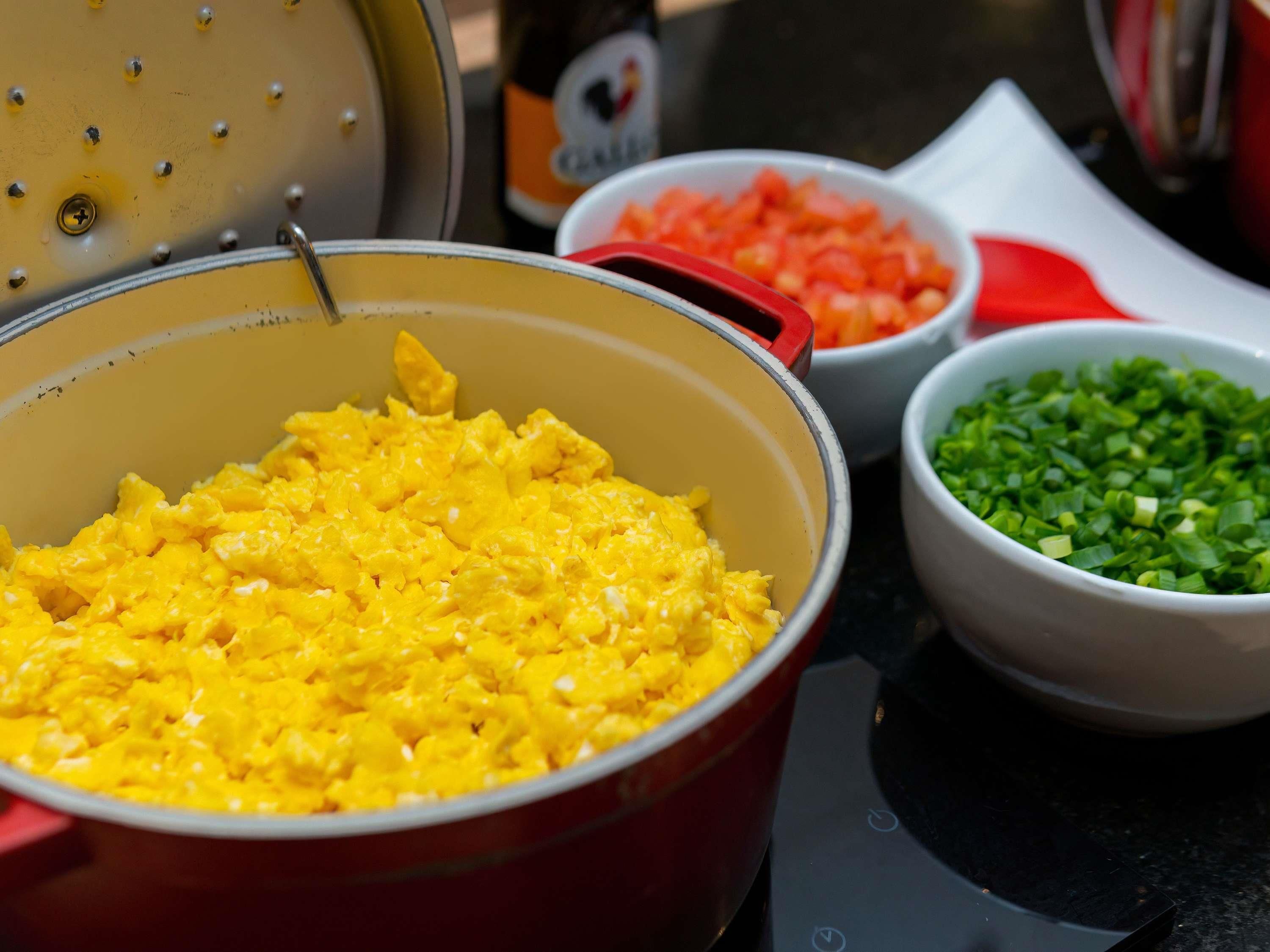 A cooking pot with scrambled eggs, a bowl of chopped carrots, and a bowl of chopped green onions on a kitchen counter.