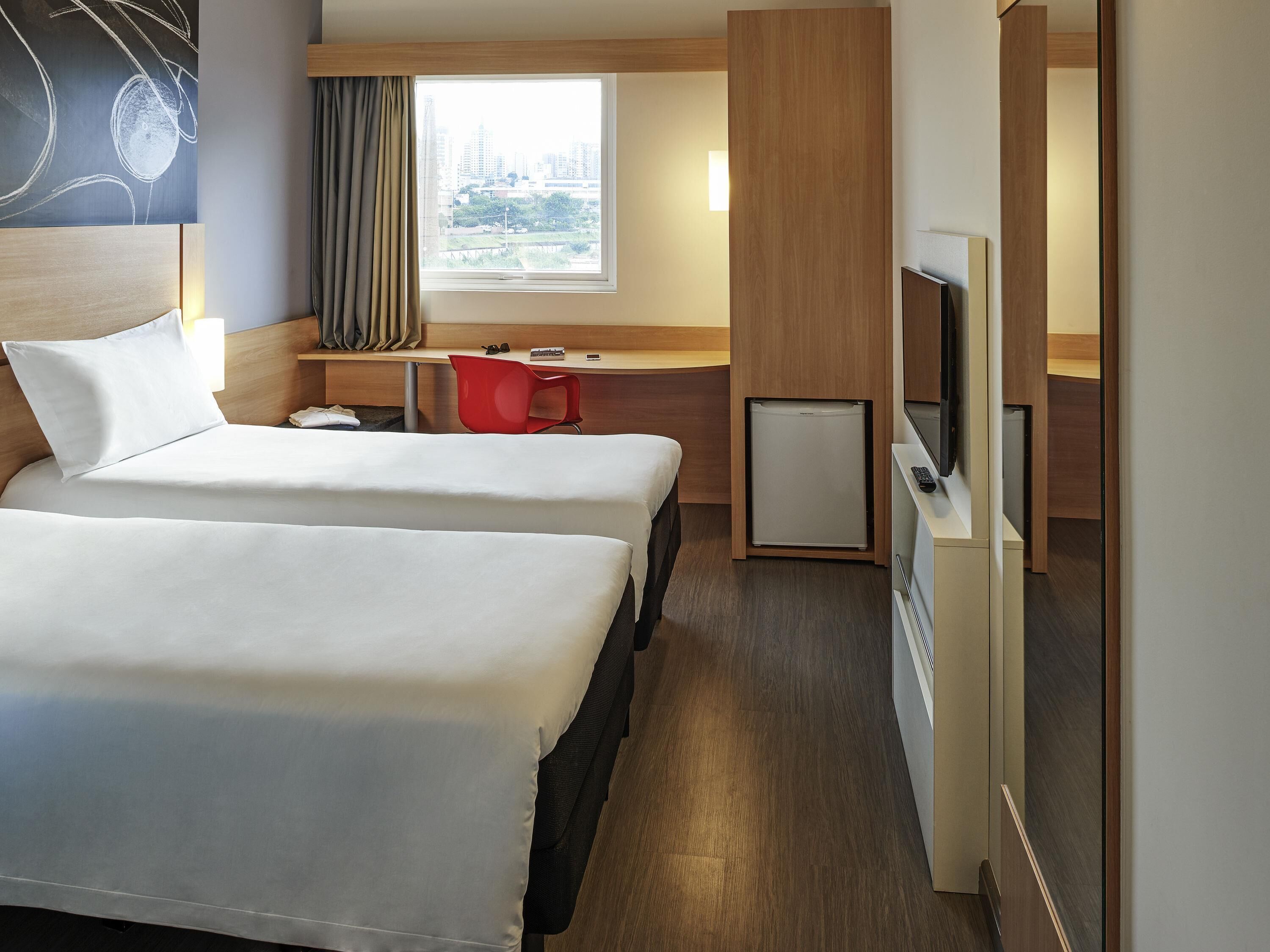 A hotel room with two beds, a desk, a chair, a TV, and a small fridge.